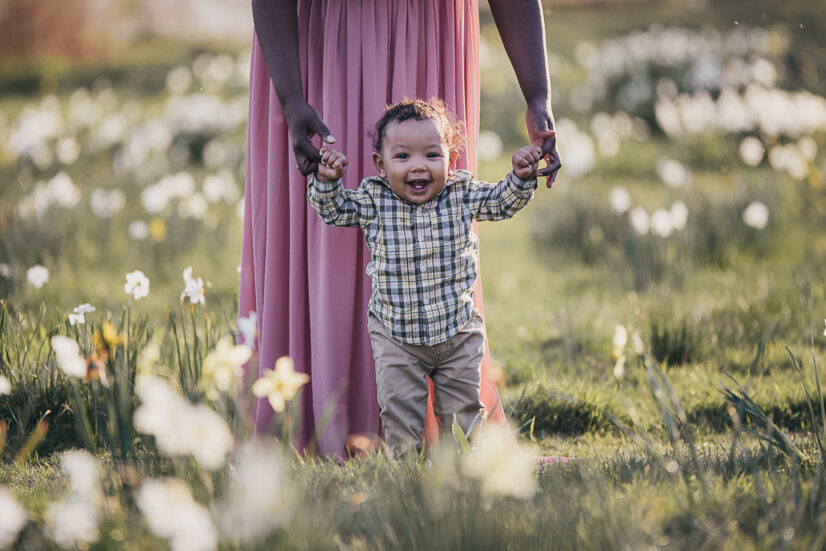 little boy in plaid shirt holding his mom's hands and laughing in a field of flowers