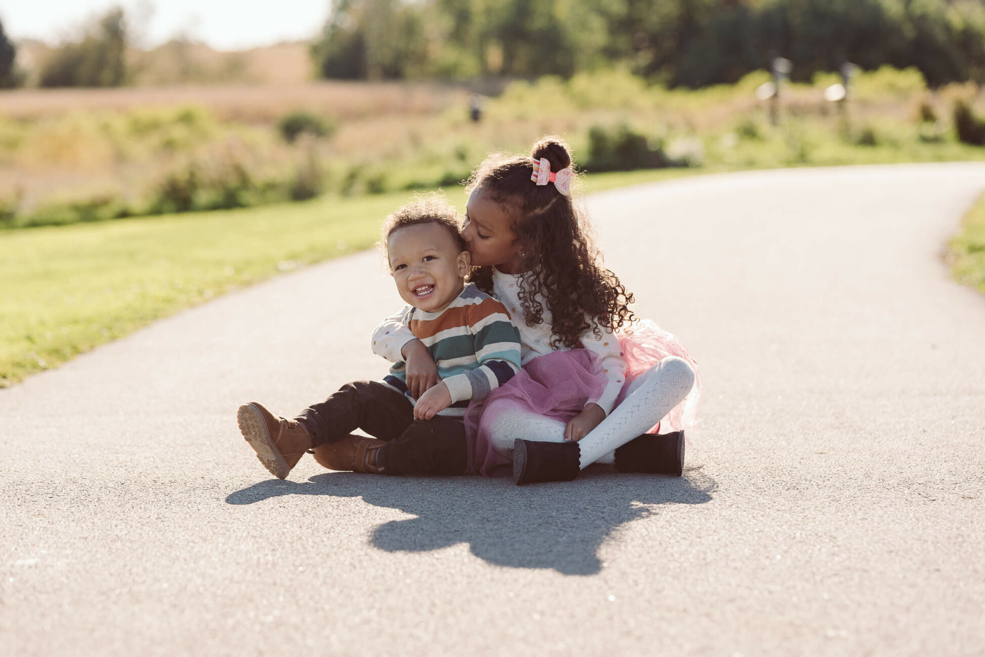 sister kissing brother while sitting on ground in the park great lakes kids apparel