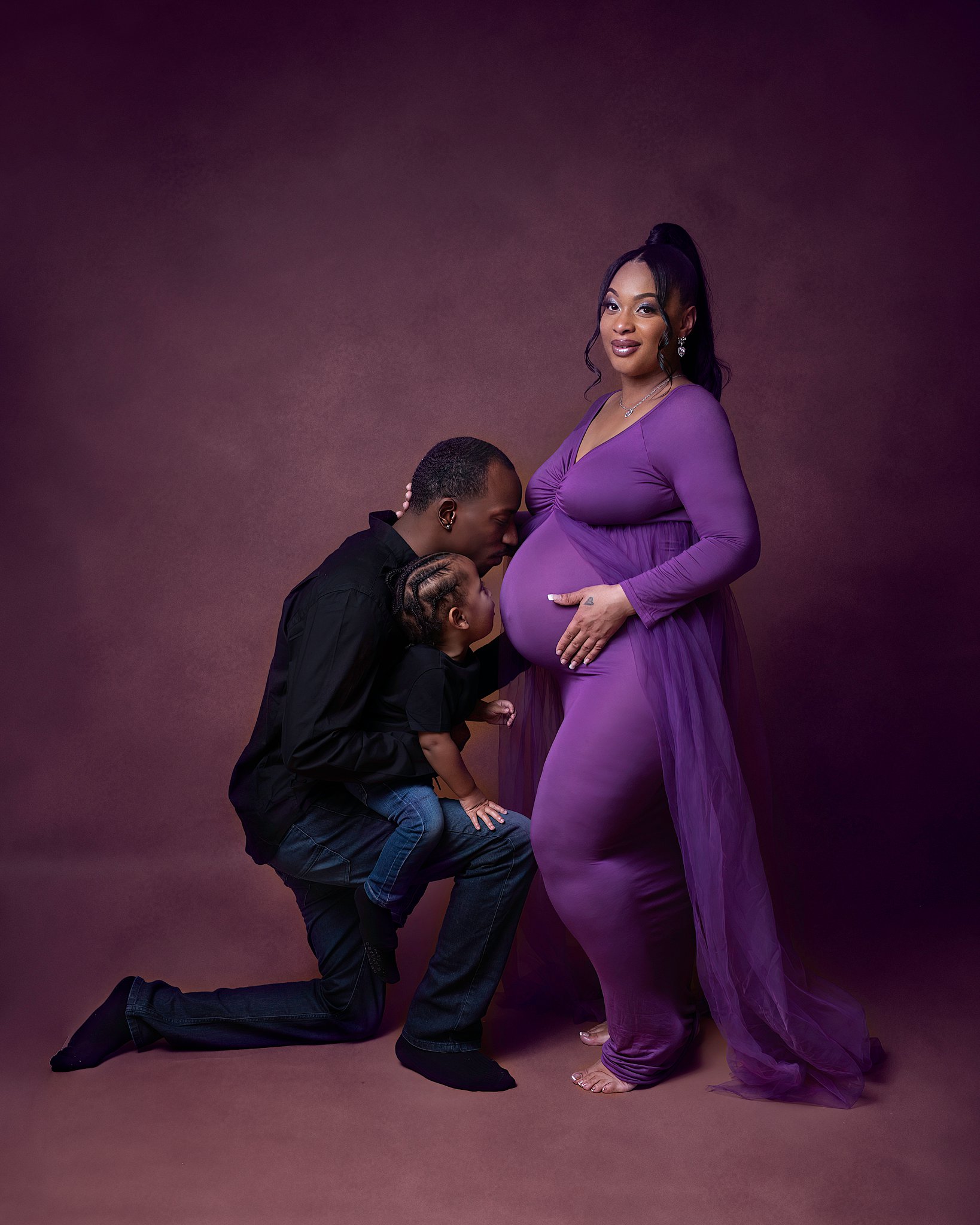 A father and his toddler son kneel in a studio, kissing the bump of his pregnant wife