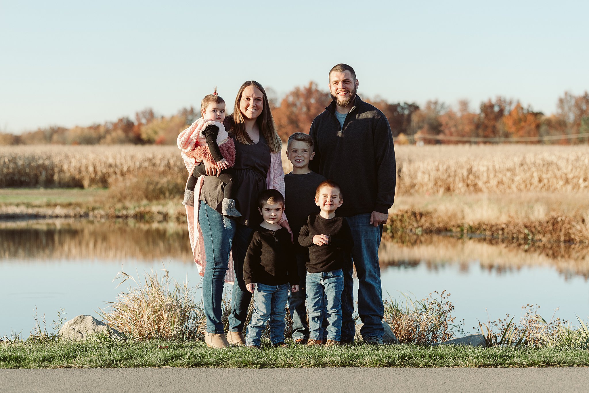 A family of six stands together in front of a lake next to a park sidewalk