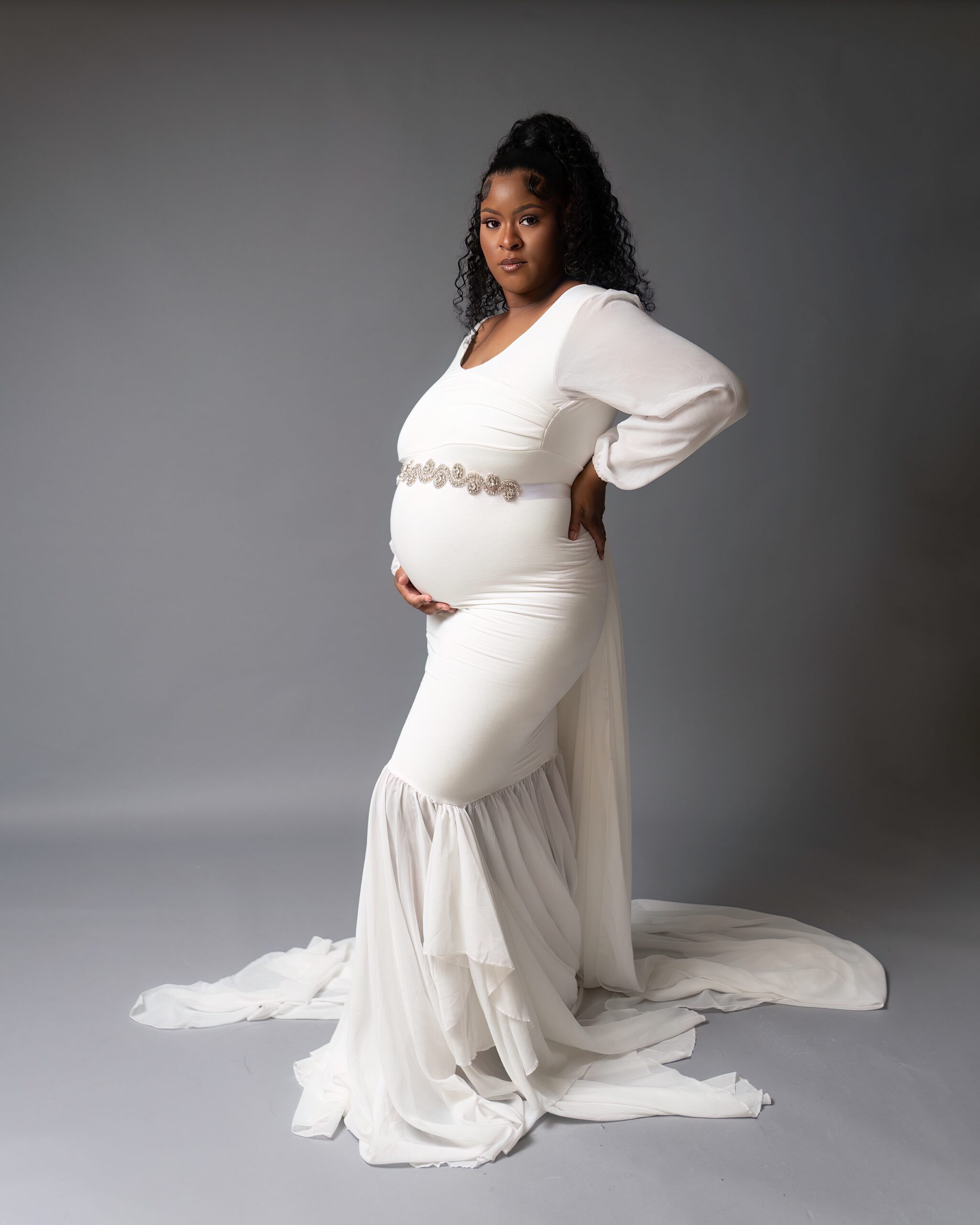 A mother to be stands in a white maternity gown with a hand on her back and other under her bump Canton Women's Center