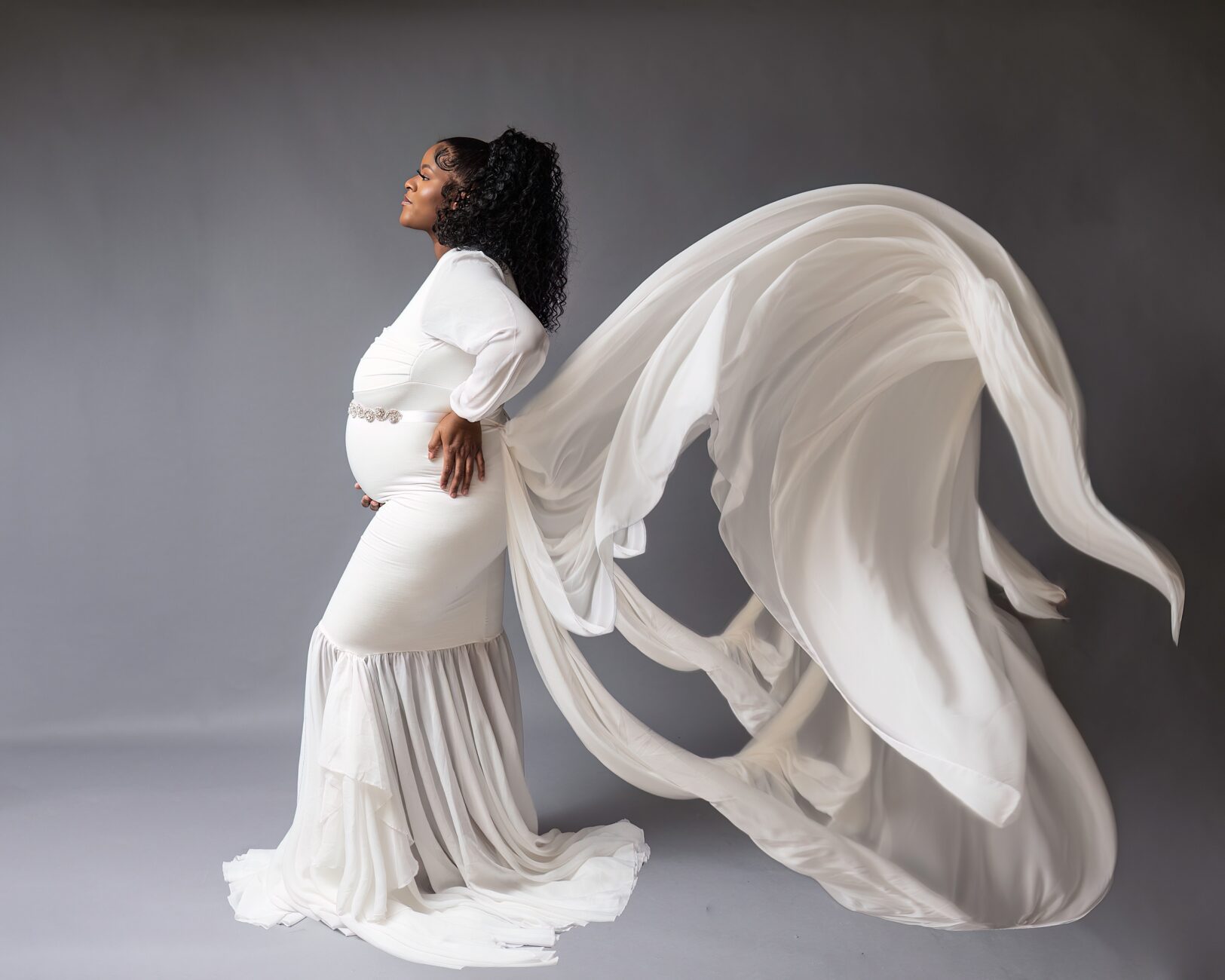 A mom to be in a white maternity gown stands in a studio with her train flowing behind her Canton Women's Center
