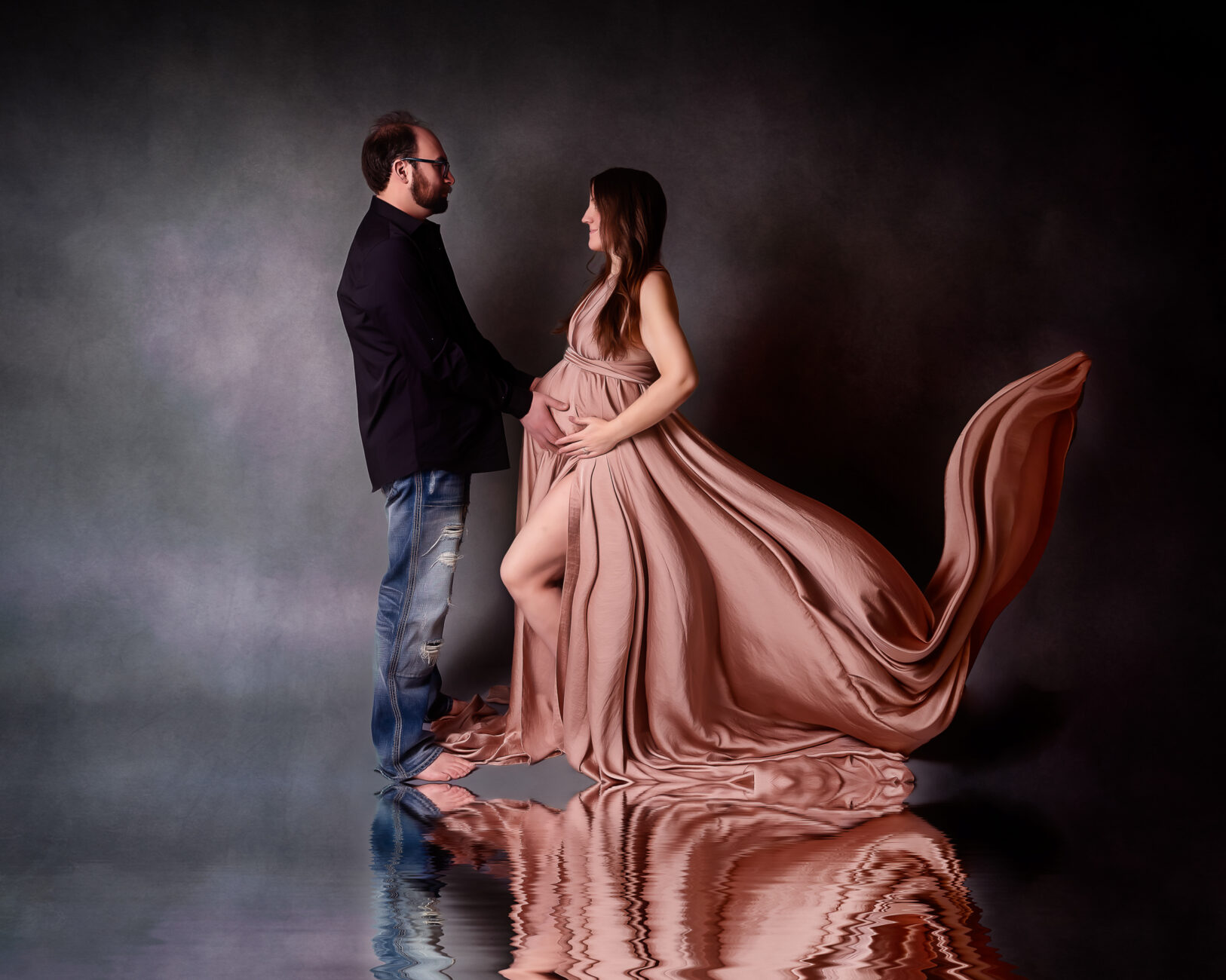 a pregnant woman and husband standing looking at each other with a pink maternity dress with reflection doulaing sisters