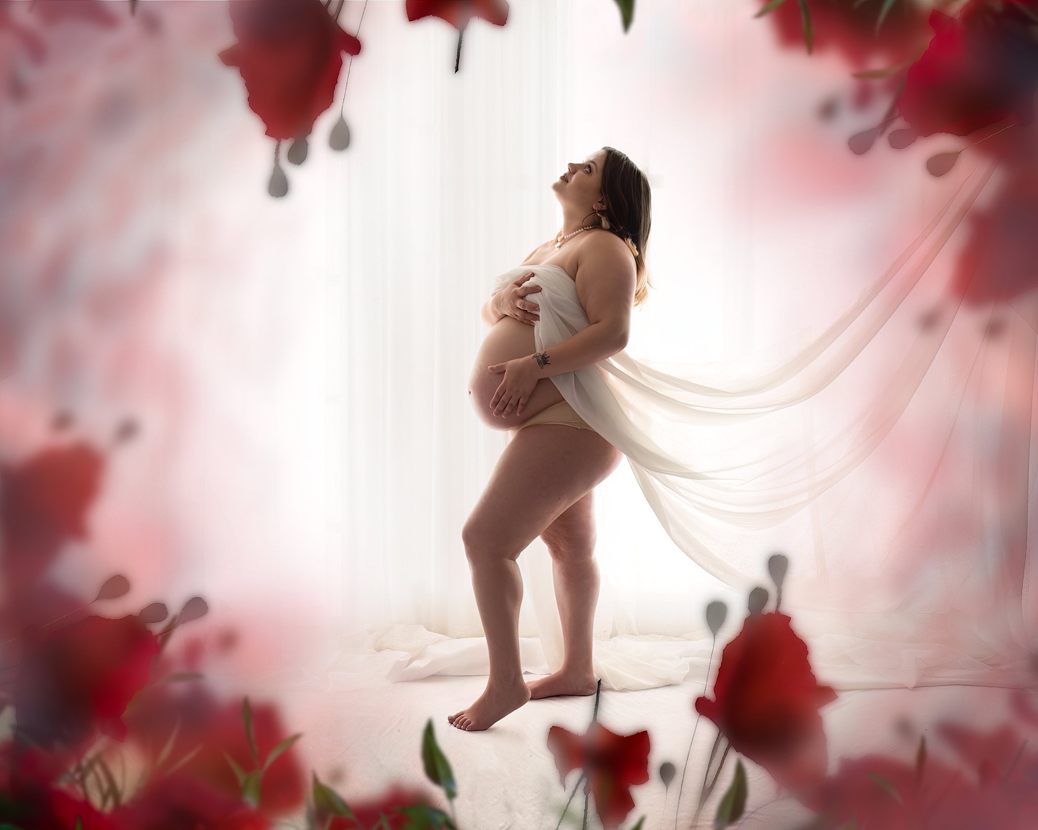 A mother to be stands in a studio in front of a window looking up in the air surrounded by red roses