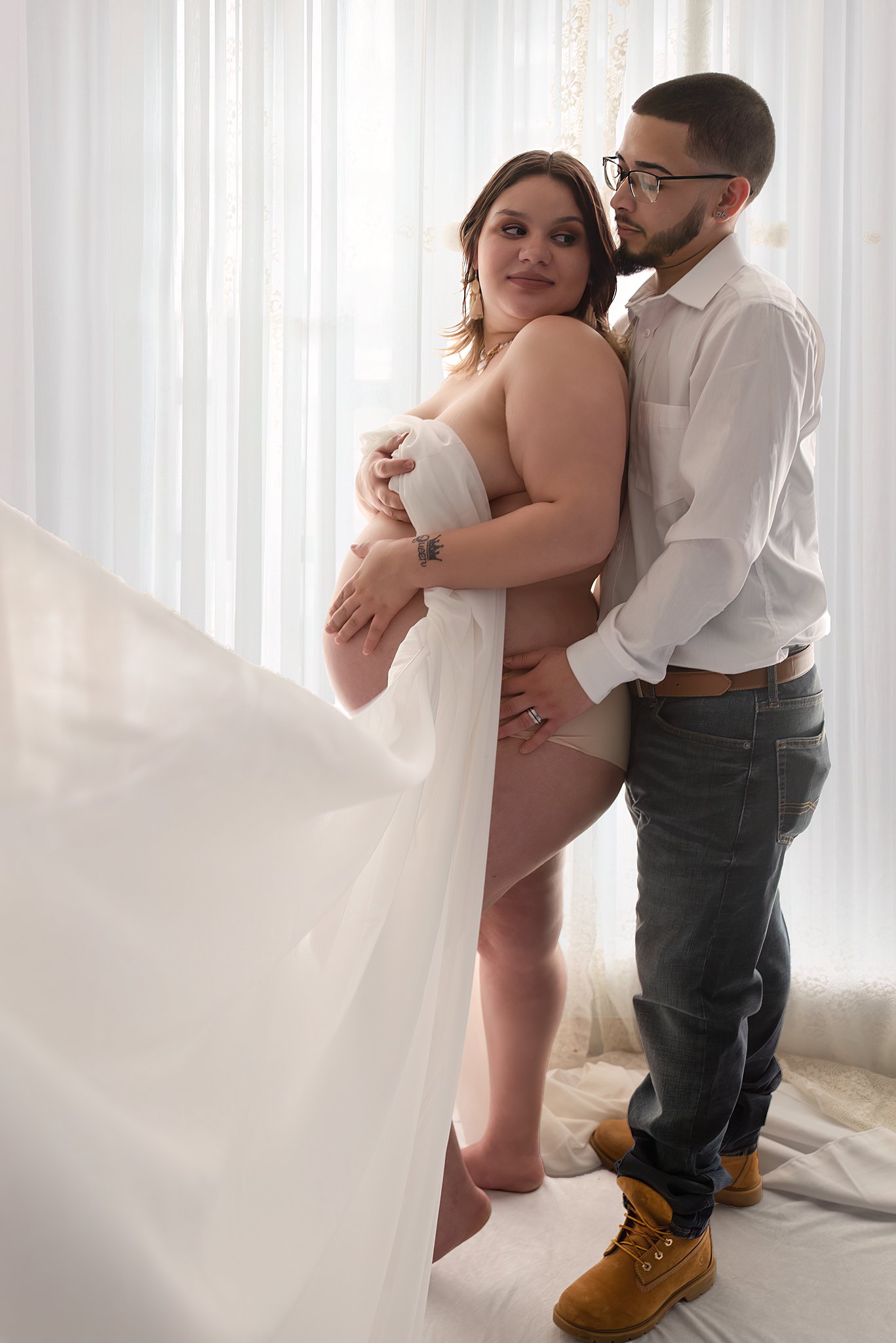 A mother to be covers herself with a white sheet while her husband hugs her from behind in front of a window nurtured foundation