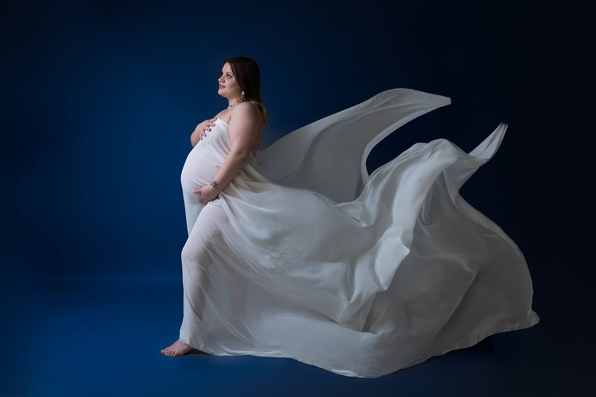 A mother to be stands in a studio holding her bump in a white maternity dress that is flowing in the wind behind her nurtured foundation