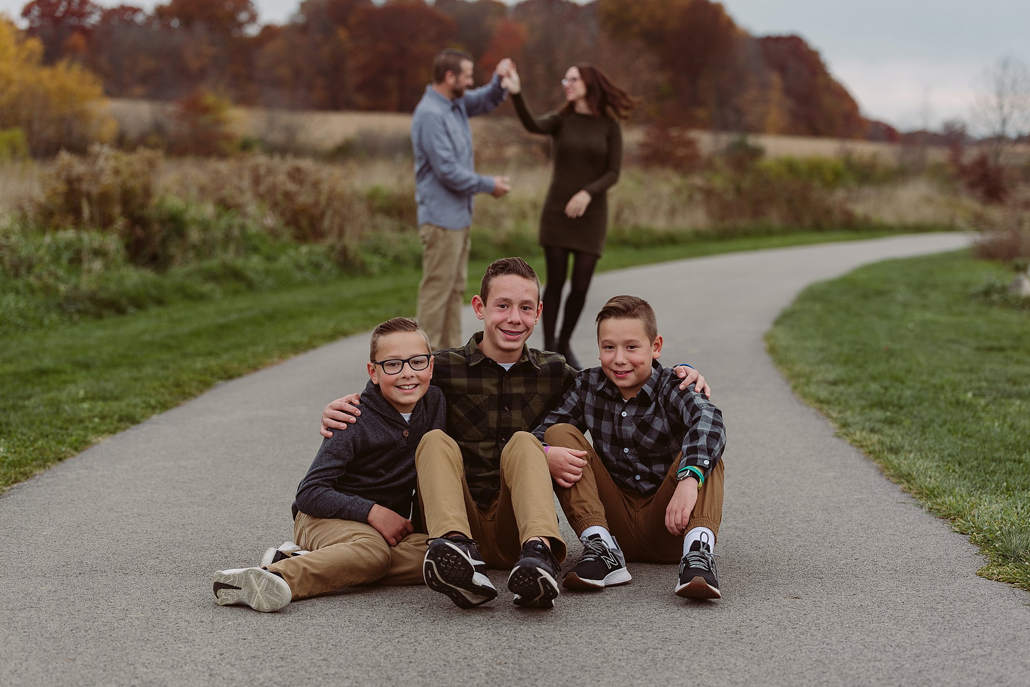 A mother and father dance together in a park path behind their three sons all sitting together