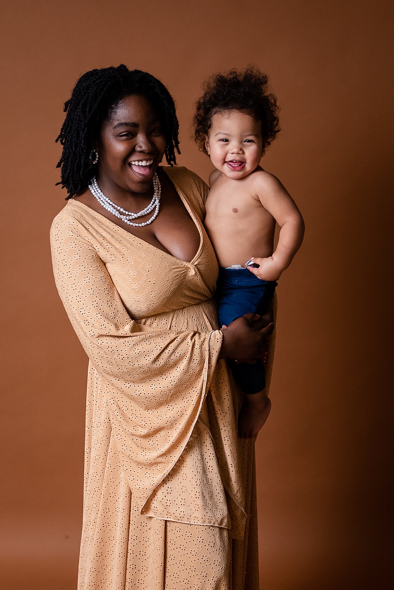 A mother in a beige dress stands in a studio holding her young toddler son on her hip playmatters toys