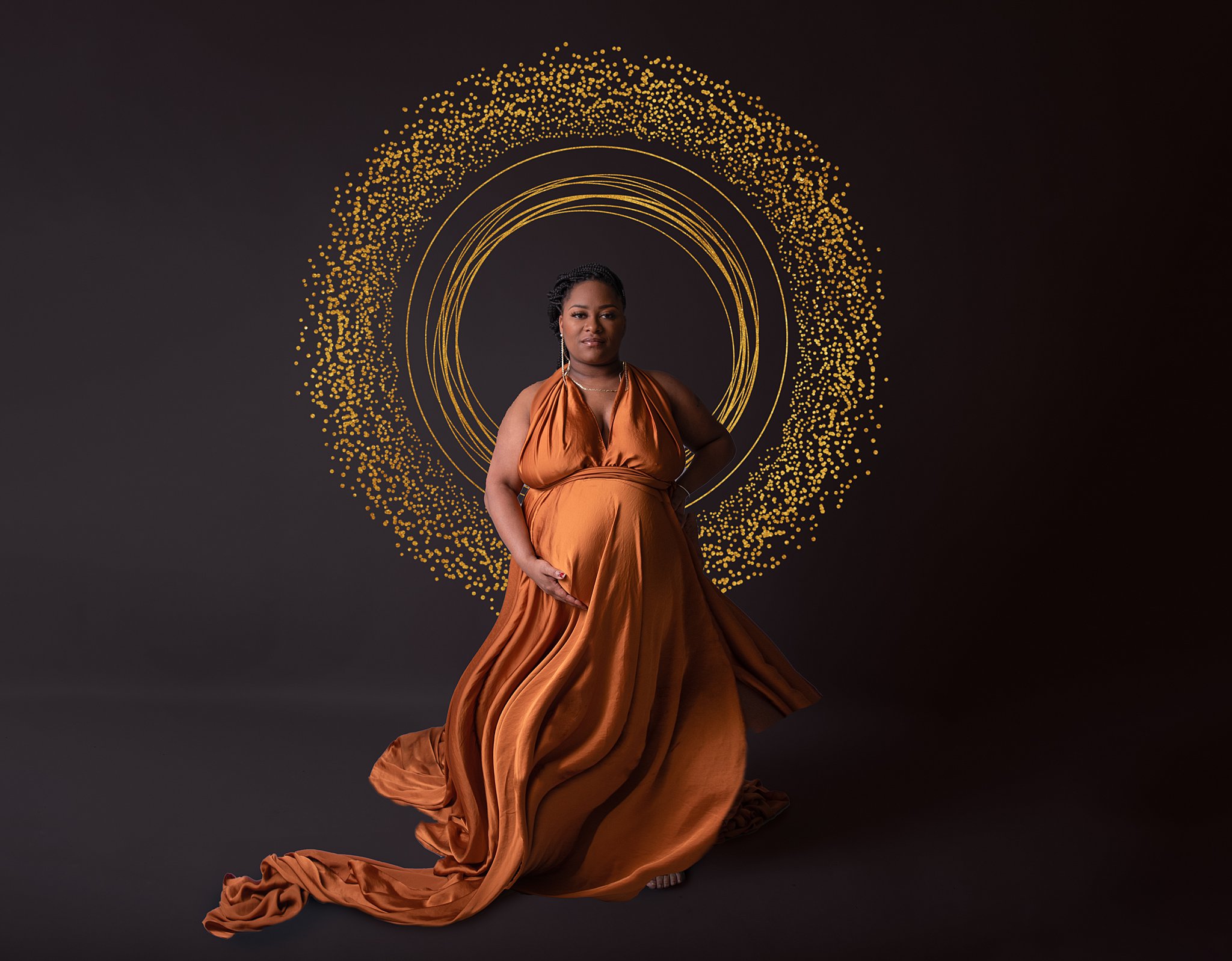 A mother to be stands in a studio wearing a long flowing orange maternity gown
