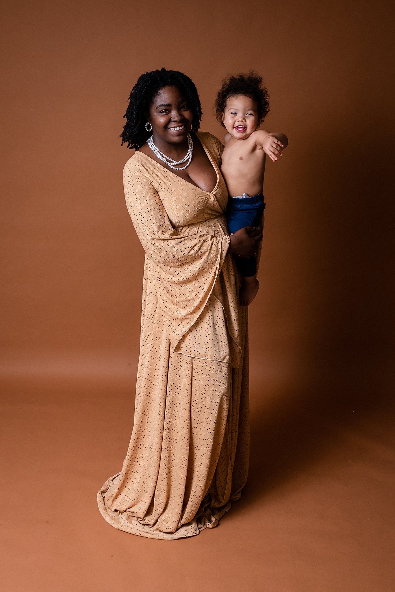 A mother stands in a studio in a tan dress holding her shirtless toddler son on her hip recess cleveland