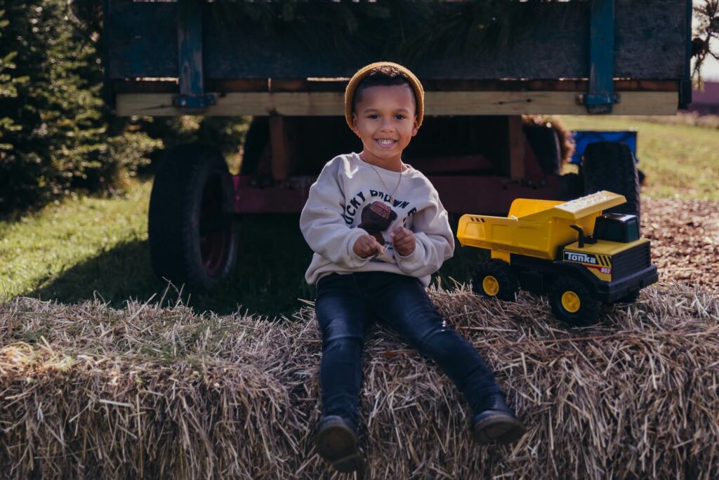 Boy sitting on haybarrel next to dump truck smiling at camera Ashton's Corner Boutique