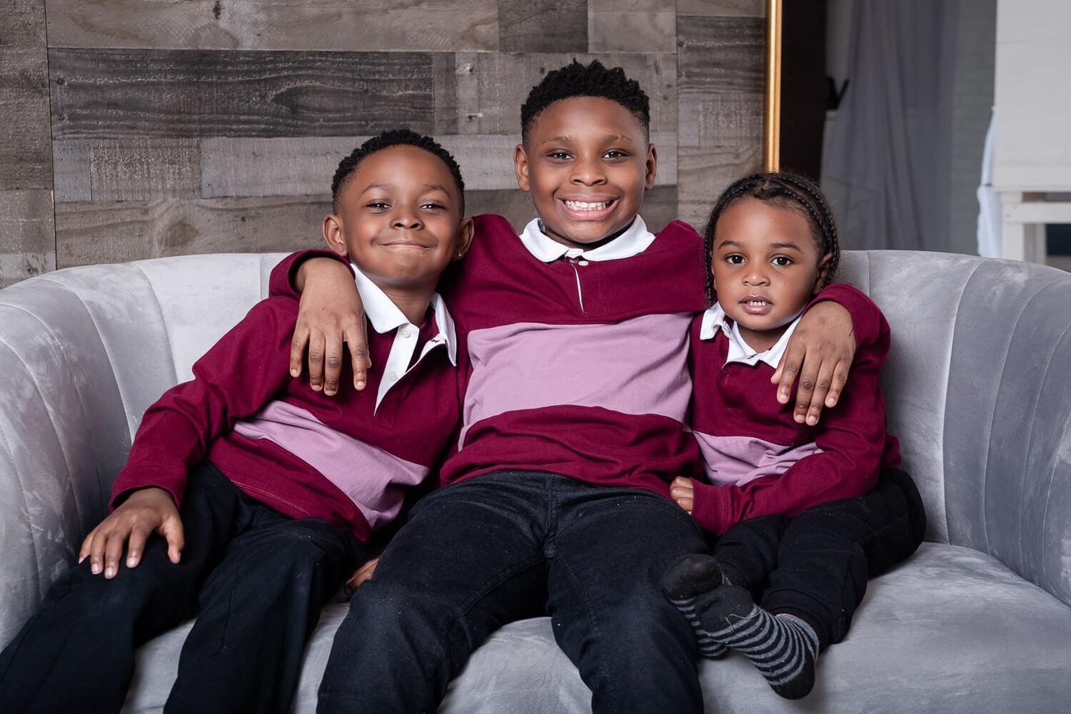 Three young boys in matching maroon and pink shirts sitting together on a gray couch, smiling and posing for a family photograph Montessori School