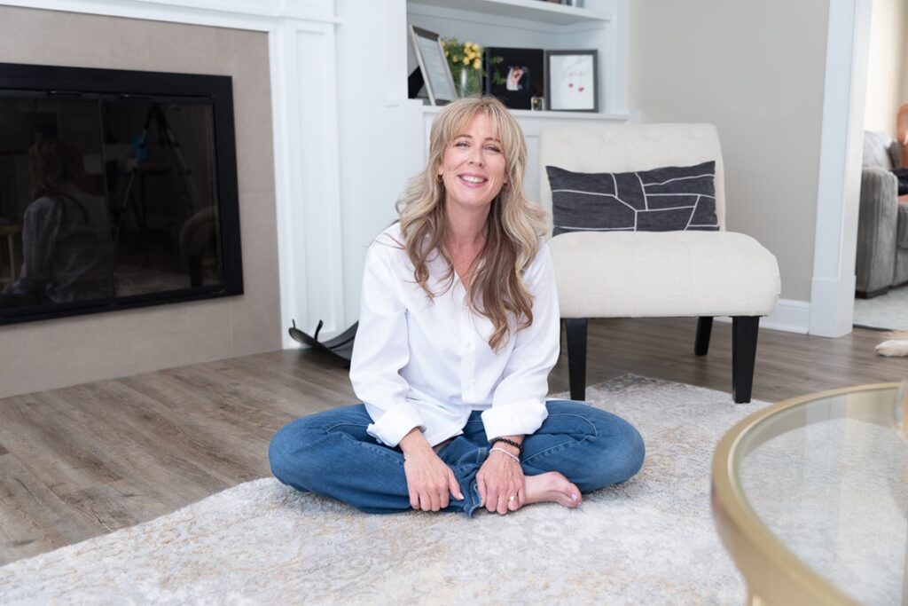 Woman in a white shirt and jeans sitting cross-legged on a cozy rug in a well-lit living room, smiling warmly at the camera. The setting is modern, with soft natural light filtering in, and a fireplace and minimalist décor visible in the background with Ivory and Birch