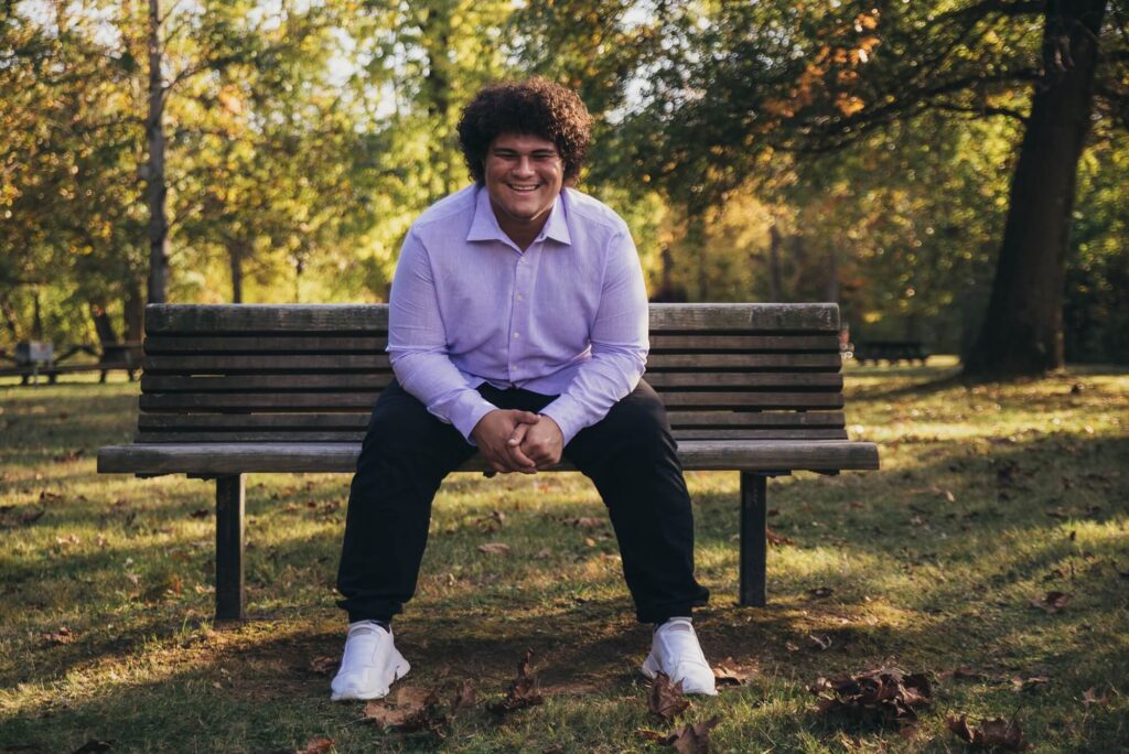 Wooster High school senior boy in park sitting on bench leaning forward smiling lookinf at camera for Acres of Fun