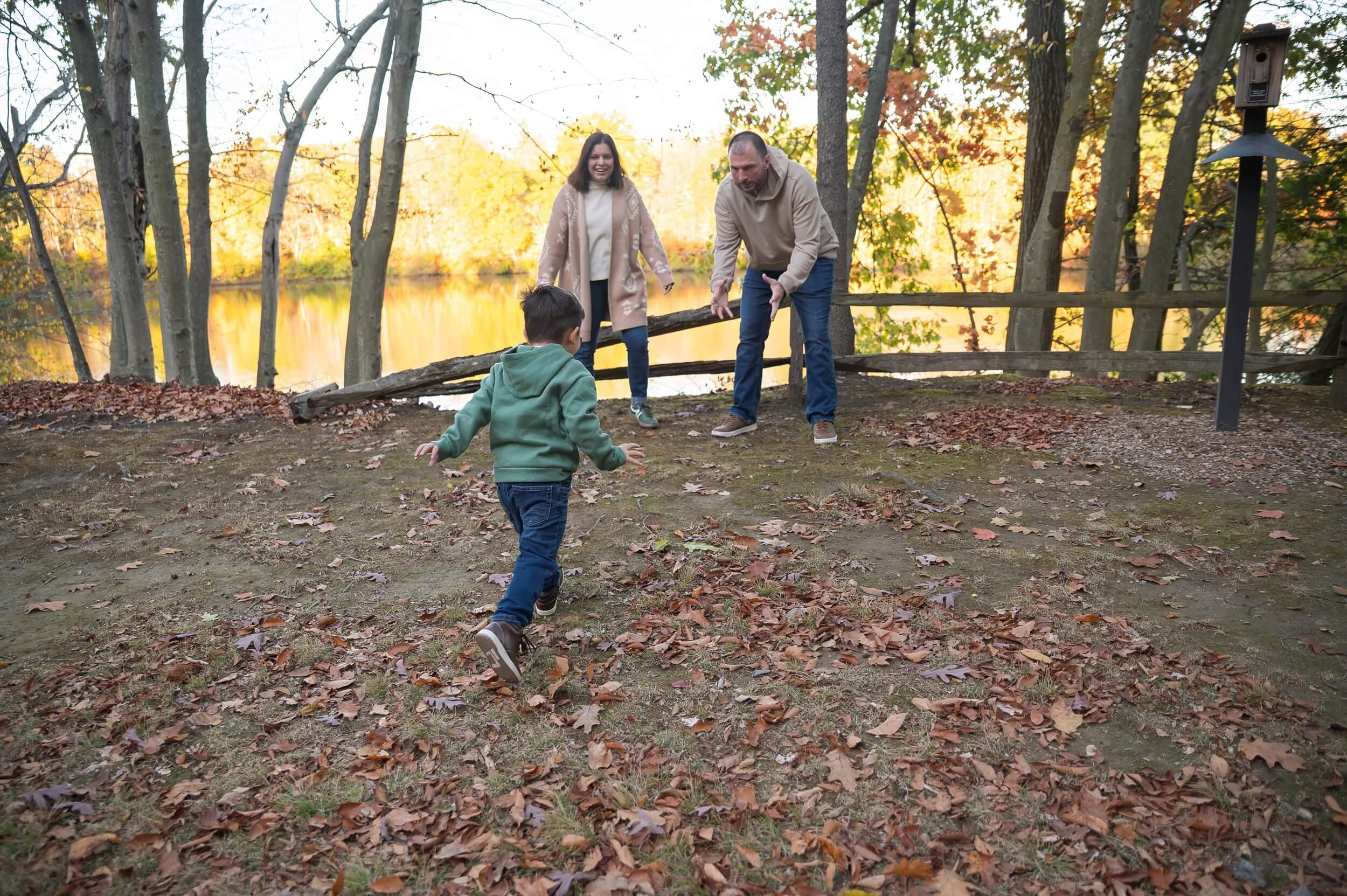 Young boy running toward his smiling parents during a fall family photo session beside a lake.