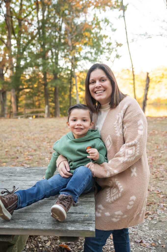 Mother smiling with her young son seated on a wooden platform in a wooded park during fall.