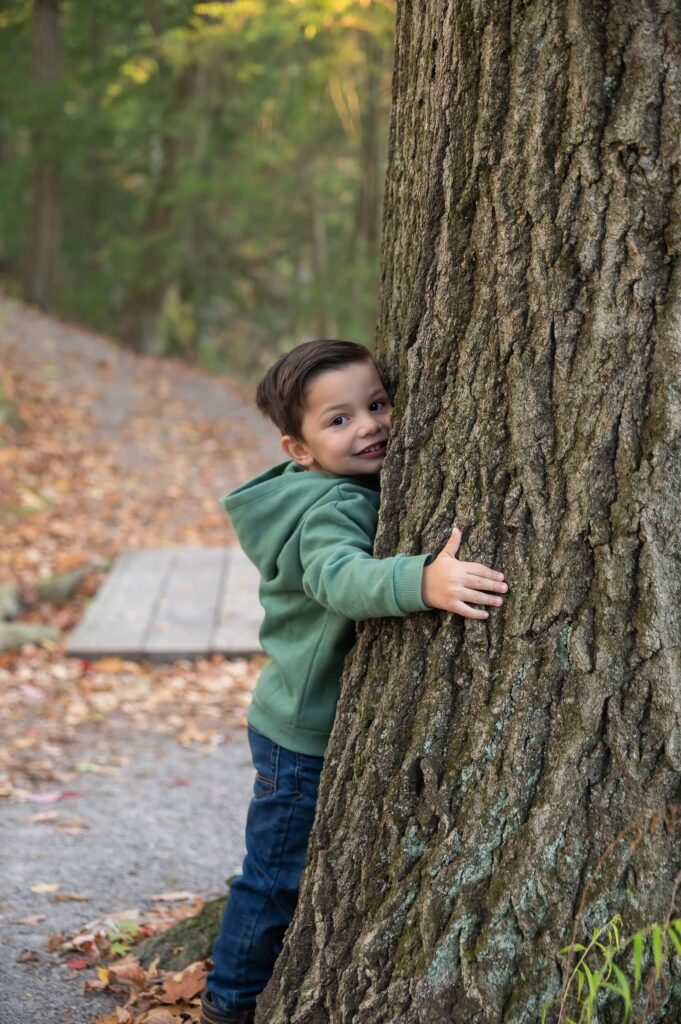 Young boy in a green hoodie smiling while hugging a large tree along a wooded trail.