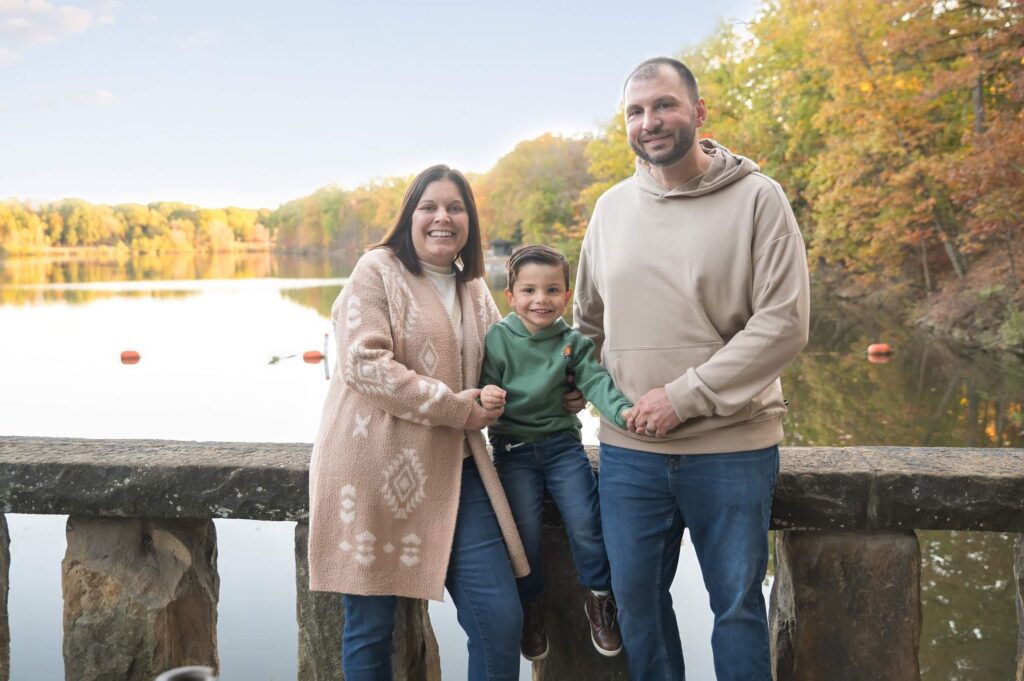 Mother, father, and young son smiling together beside a peaceful lake surrounded by colorful fall trees for Sensory-Friendly Family Events