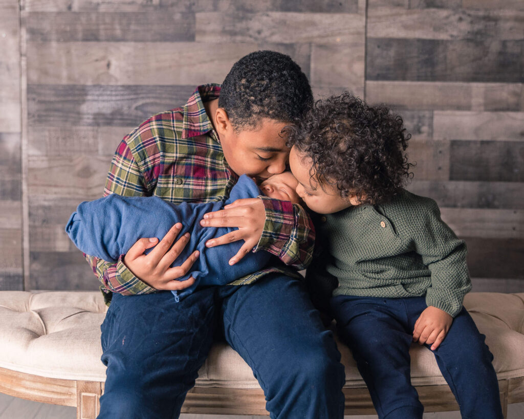Two brothers gently kissing their swaddled newborn sibling during a cozy portrait session.