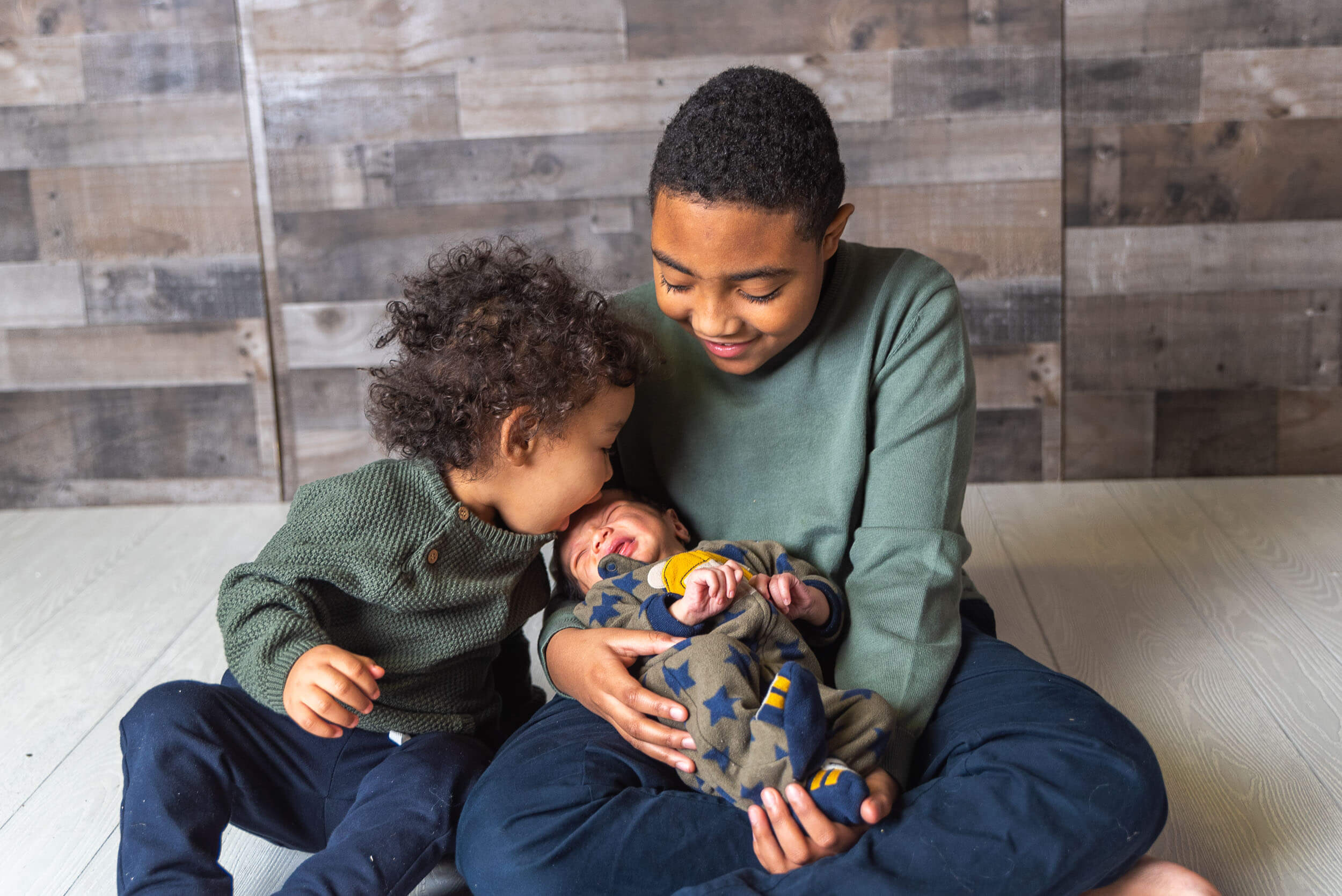 Two older brothers smiling and looking down at their newborn sibling during an indoor family photo session.